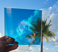 Hand holding a transparent automotive window tint film sample reflecting a blue sky and palm tree against a clear outdoor sky. The reflective tint demonstrates its subtle appearance and light-reflective finish for custom vehicle styling.