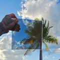 Hyper chrome automotive window tint sample with a highly reflective mirror-like finish, held outdoors against a bright blue sky with palm trees reflected across the surface. Hyper chrome tint showcasing its vibrant colour, reflective appearance, and distinctive custom vehicle styling potential.