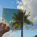 Pure gold chrome automotive window tint sample with a highly reflective mirror-like finish, held outdoors against a bright blue sky with palm trees reflected across the surface. Pure gold chrome tint showcasing its vibrant colour, reflective appearance, and distinctive custom vehicle styling potential.
