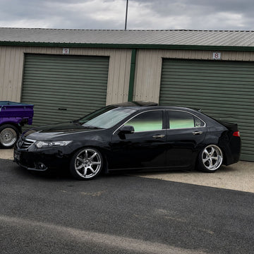 Black customized sedan featuring aftermarket silver multi-spoke wheels, parked in front of a beige and green garage building. Modified vehicle showcasing clean custom automotive styling and performance-inspired design.