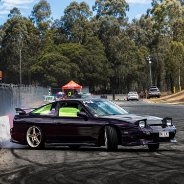 Dark purple customized sports car drifting on an asphalt track, with trees and a race event setup in the background. Custom drift car styling featuring automotive window tint and styling film.