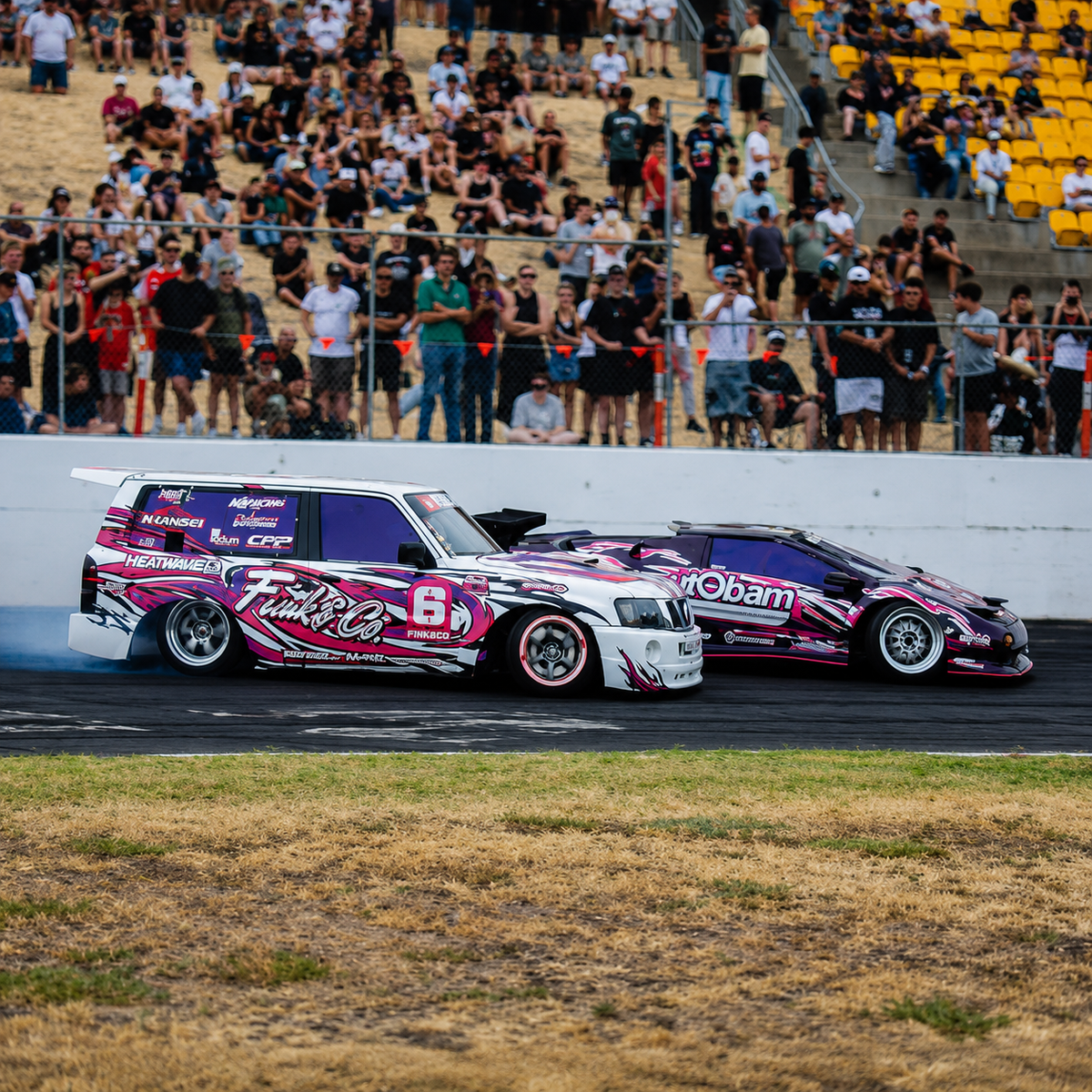 Two race cars competing on a track in front of a large crowd, with a white car featuring pink and purple graphics and number 6 leading a purple car with pink and white graphics. Automotive racing styling and custom car graphics.