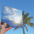 Steel blue chrome automotive window tint sample with a highly reflective mirror-like finish, held outdoors against a bright blue sky with palm trees reflected across the surface. Steel blue chrome tint showcasing its vibrant colour, reflective appearance, and distinctive custom vehicle styling potential.
