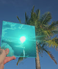Turquoise chrome automotive window tint sample with a highly reflective finish, held outdoors against a bright blue sky and palm trees. The turquoise chrome tint reflects sunlight and surrounding scenery, showcasing its vibrant colour and mirror-like appearance for custom vehicle styling.