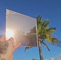 Brazen bonze chrome automotive window tint sample with a highly reflective mirror-like finish, held outdoors against a bright blue sky with palm trees reflected across the surface. Brazen bronzechrome tint showcasing its vibrant colour, reflective appearance, and distinctive custom vehicle styling potential.