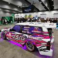 Heavily customized white Ford Ranger featuring a vibrant pink, black, and white flame wrap, custom wheels, and purple underglow, displayed at a car show with a crowd in the background. Custom automotive styling and vehicle wrap design.
