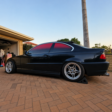 Lowered black BMW coupe featuring tinted windows and silver multi-spoke wheels, parked on a brick driveway in front of a residential garage. Custom automotive styling with aftermarket wheels and automotive window tint.