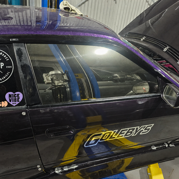 Close-up of a black car door featuring a purple glitter styling line along the window trim, a visible “GOLEBYS” decal, and a reflective tinted side window showing the garage interior. Custom automotive window styling and reflective window tint.