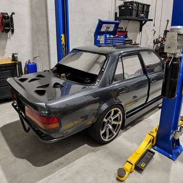Dark gray custom-modified sedan featuring a large rear trunk vent design, parked inside a professional garage workshop between blue vehicle lifts. Custom automotive styling and performance-inspired vehicle design.